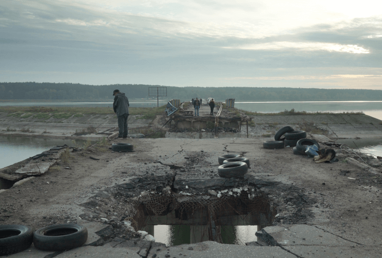 Grupa ludzi stoi na dalekim skraju zniszczonego, rozpadającego się mostu nad zbiornikiem wodnym. Opony i gruz są porozrzucane na zniszczonej konstrukcji, z dużą dziurą pośrodku. Niebo jest zachmurzone.