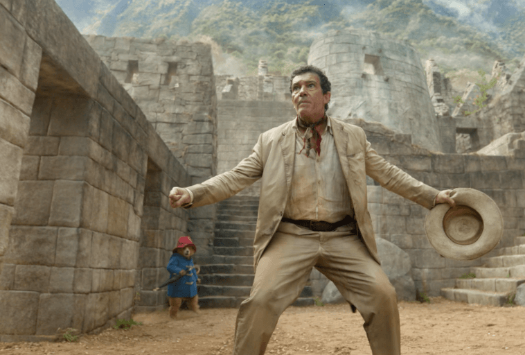 Mężczyzna w beżowym garniturze stoi wyzywająco trzymając kapelusz, za nim widać starożytne kamienne ruiny i góry. W tle na schodach stoi mały niedźwiedź w niebieskim płaszczu i czerwonym kapeluszu.