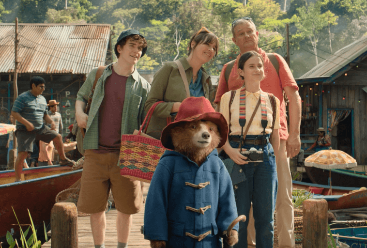 Brązowy niedźwiedź w niebieskim płaszczu i czerwonym kapeluszu stoi na drewnianym pomoście przed czterema uśmiechniętymi ludźmi, w tle widać łodzie, kolorowe parasole i rustykalne budynki.