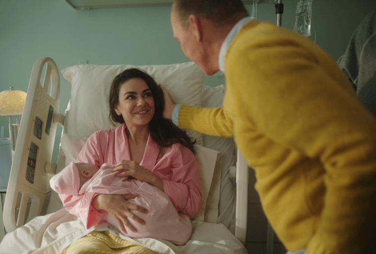Kobieta leżąca na szpitalnym łóżku, ubrana w różowy szlafrok, uśmiecha się do mężczyzny w żółtym swetrze, trzymając na rękach noworodka owiniętego w różowy kocyk.