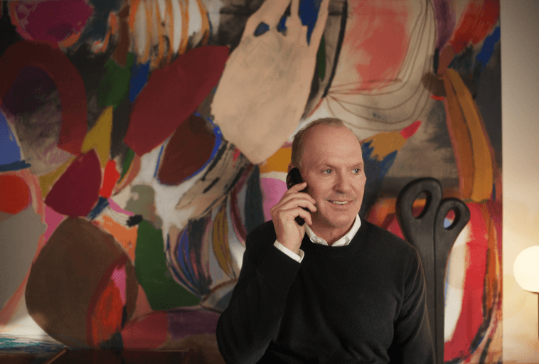 Mężczyzna w ciemnym swetrze uśmiecha się, rozmawiając przez telefon, siedząc przed kolorowym abstrakcyjnym obrazem o odważnych kształtach i pociągnięciach pędzla. Obok niego widać rzeźbiarskie krzesło i okrągłą lampę.