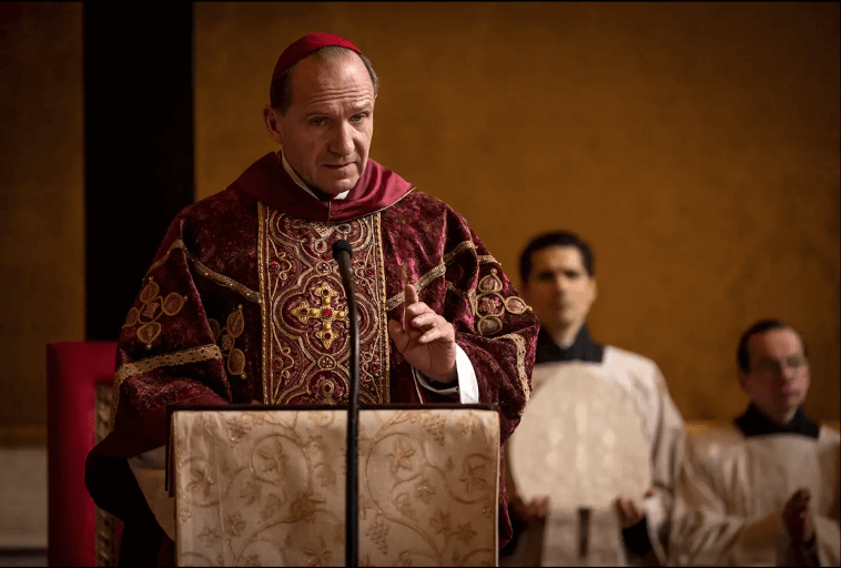 Biskup w ozdobnych bordowych i złotych szatach przemawia z ambony, gestykulując jedną ręką. W tle stoją dwaj księża w białych szatach, jeden trzyma okrągły przedmiot. Sceną jest kościół ze złotymi ścianami.