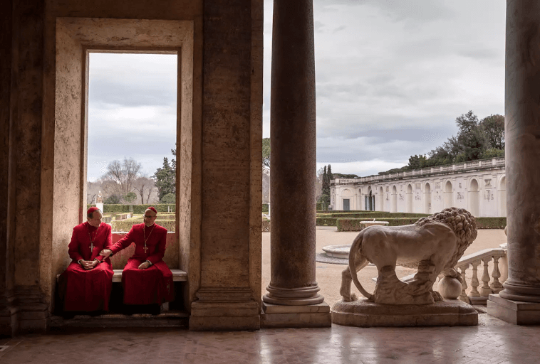Dwie osoby w czerwonych szatach siedzą na ławce otoczonej wysokimi kamiennymi kolumnami. Po prawej stronie znajduje się biała statua lwa, a w tle, pod zachmurzonym niebem, widać ozdobny budynek i ogród.