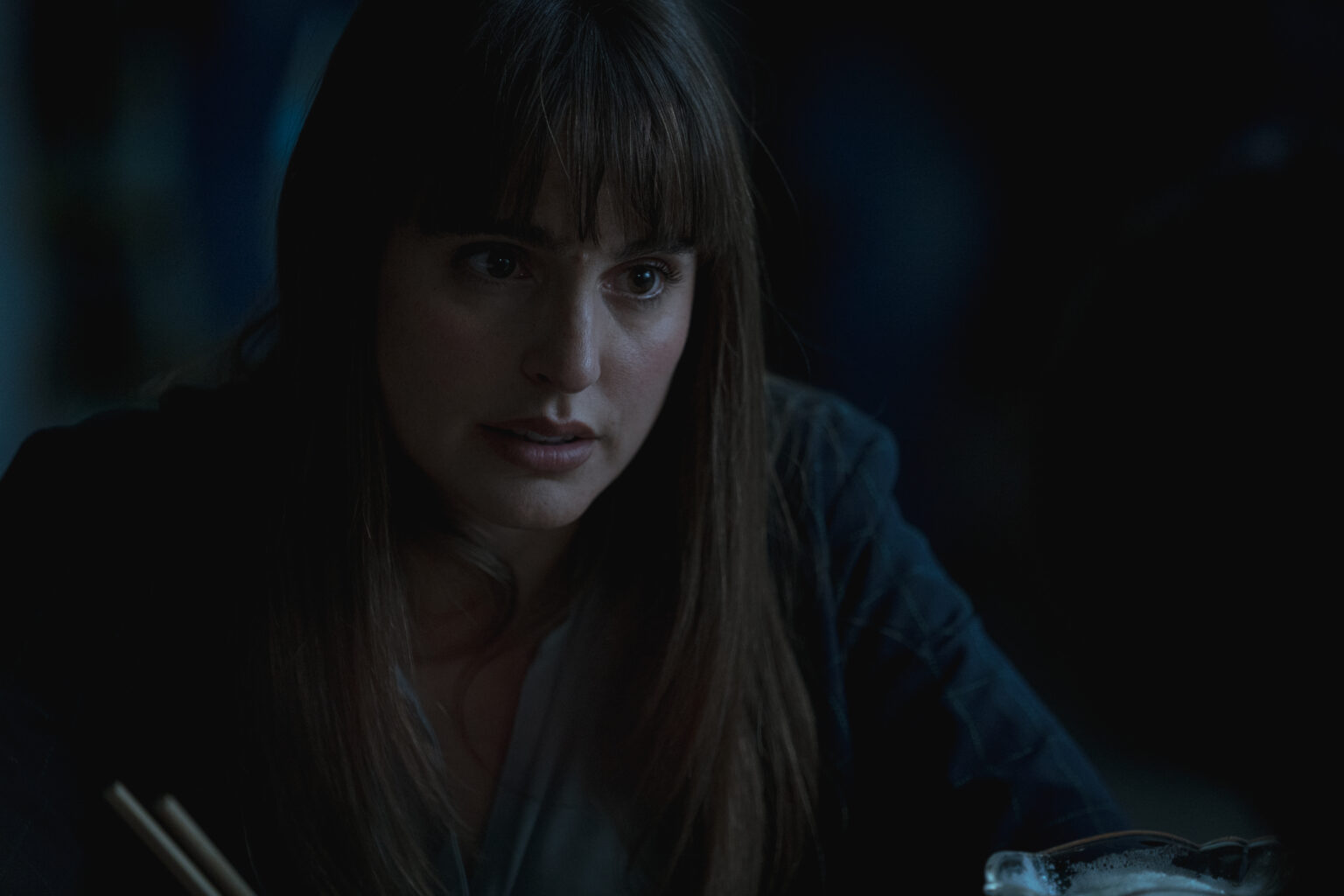 Kobieta o długich brązowych włosach i grzywce uważnie patrzy przed siebie w słabo oświetlonym otoczeniu. W jej dłoni widać pałeczki, a na pierwszym planie znajduje się zamglona szklanka.