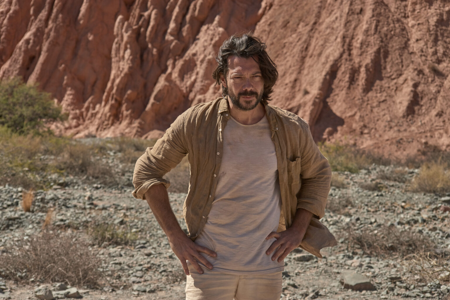 Mężczyzna z potarganymi ciemnymi włosami i brodą stoi na zewnątrz przed rudawymi skalistymi klifami, ubrany w opaloną koszulę na beżowym T-shircie i beżowe spodnie, z rękami na biodrach. Ziemia jest skalista z rzadką roślinnością.
