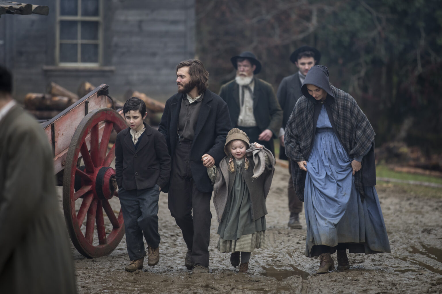 Grupa ludzi w staromodnych ubraniach idzie błotnistą drogą w pobliżu drewnianego wozu. Mężczyzna trzyma za ręce dwójkę dzieci, a kobieta w niebieskiej sukience i szalu idzie obok nich. W tle widać innych ludzi i budynki.