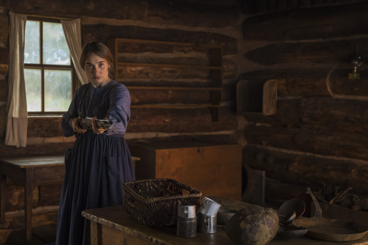 Kobieta w niebieskiej sukience stoi w słabo oświetlonej chacie z bali, trzymając karabin skierowany do przodu. Wygląda na czujną i zdeterminowaną. Wokół niej widać rustykalne drewniane meble i kosze. Światło wpada przez okno za nią.