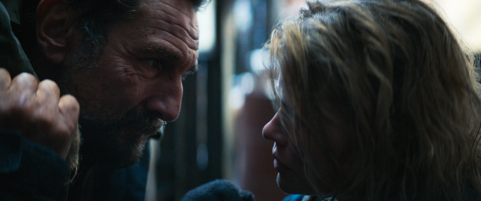 Brodaty mężczyzna i kobieta z potarganymi blond włosami stoją naprzeciw siebie, oboje z intensywnymi, poważnymi wyrazami twarzy w słabo oświetlonym otoczeniu.