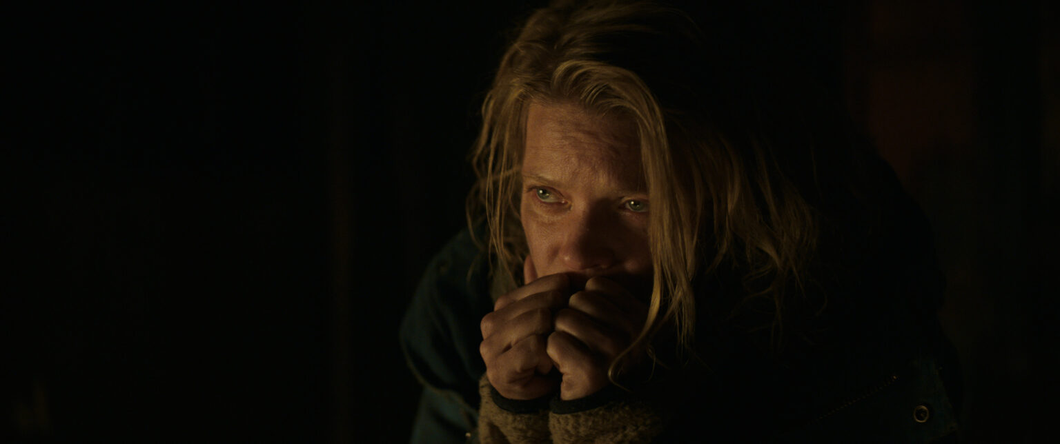 Osoba z długimi, jasnymi włosami wygląda na zaniepokojoną, trzymając ręce blisko ust, aby się ogrzać lub poczuć komfort, w słabym oświetleniu. Nosi ciemny płaszcz z podszewką z brązowego polaru.