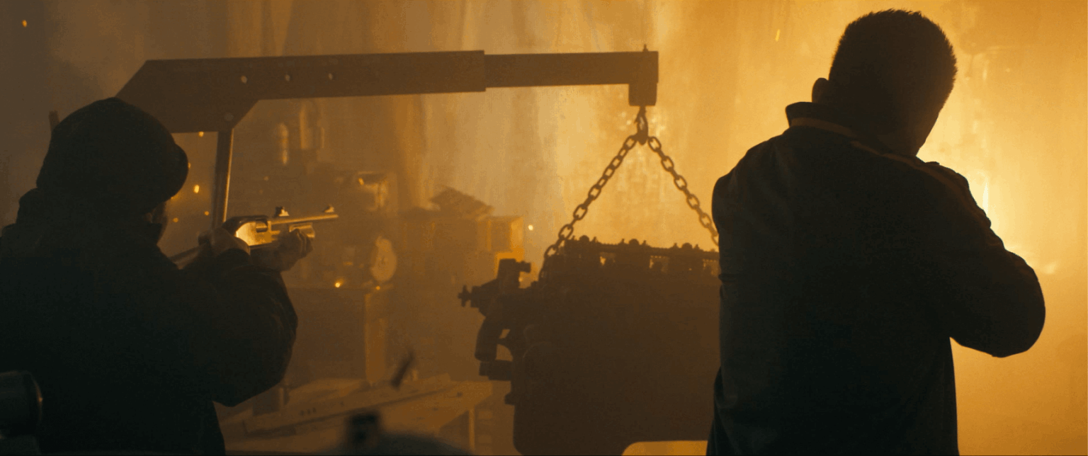Dwie mroczne postacie stoją w przyćmionym, oświetlonym na pomarańczowo przemysłowym otoczeniu, jedna celuje z broni, a druga jest odwrócona. W tle widać ciężkie maszyny i łańcuchy, co potęguje napiętą atmosferę.