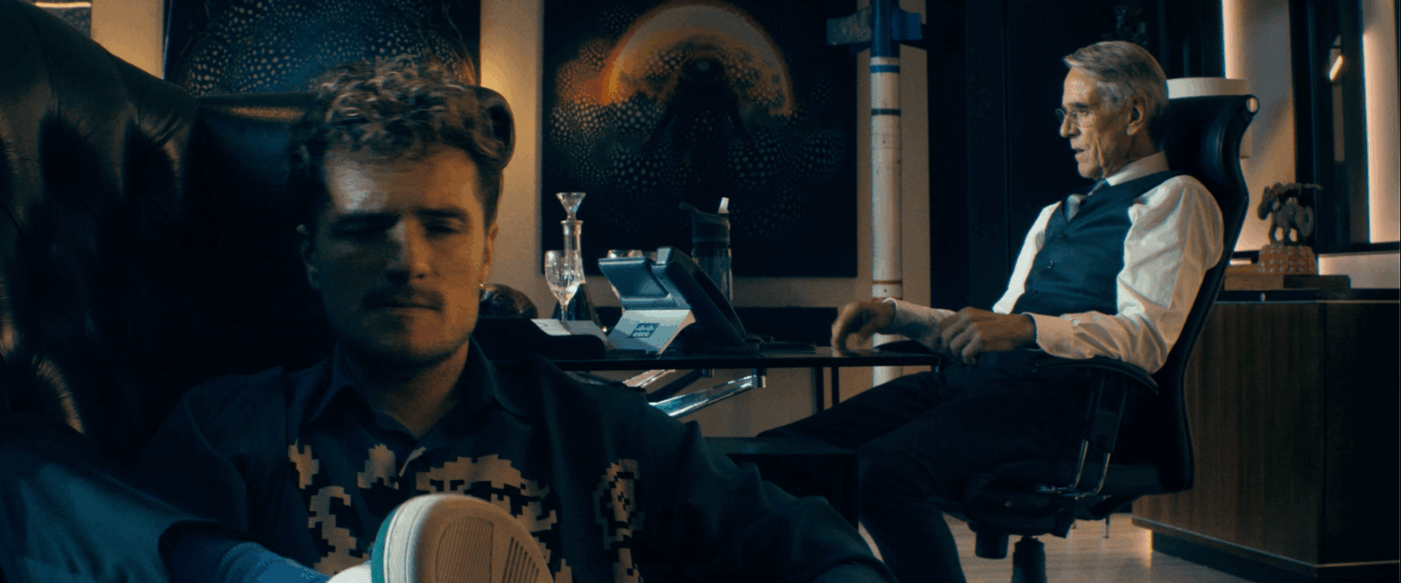 Mężczyzna z kręconymi włosami siedzi swobodnie na pierwszym planie, z zamkniętymi oczami, podczas gdy starszy mężczyzna w kamizelce siedzi przy biurku w tle, pracując na komputerze. Na ścianach za nimi wiszą abstrakcyjne dzieła sztuki.