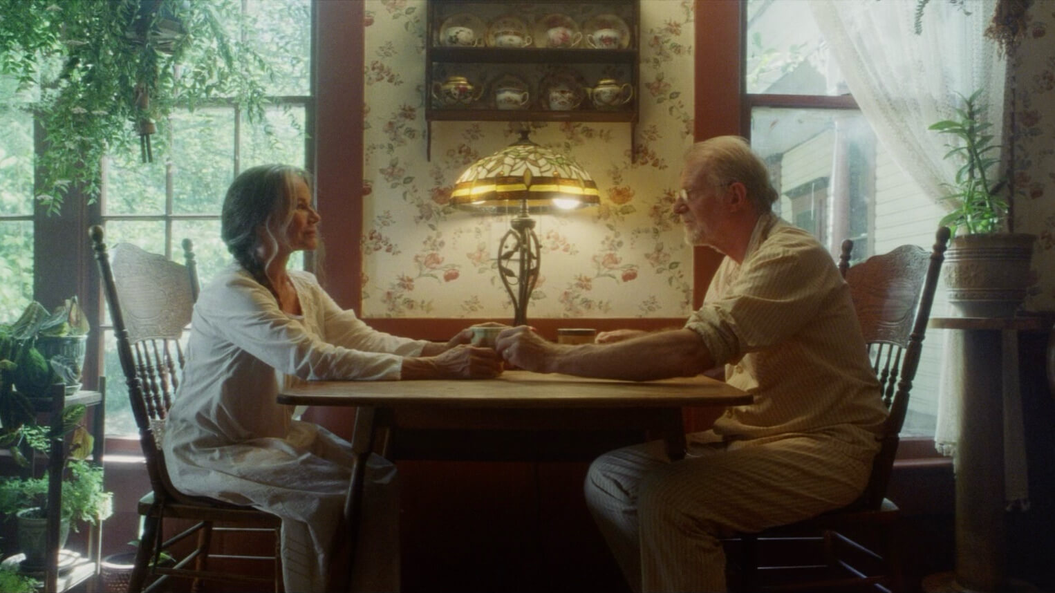 Starsza para siedzi przy drewnianym stole w przytulnej, słonecznej kuchni, trzymając się za ręce i uśmiechając się do siebie. Rośliny, kwiatowa tapeta i witrażowa lampa tworzą ciepłą, zachęcającą atmosferę.