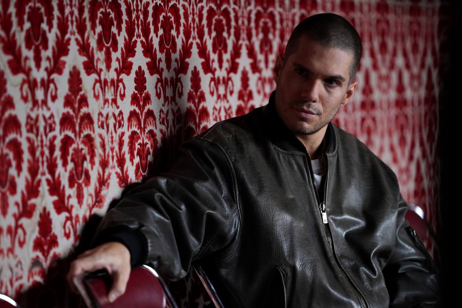 Mężczyzna z krótkimi włosami, ubrany w ciemną skórzaną kurtkę, siedzi na krześle na tle czerwono-białej, wzorzystej tapety, patrząc w bok z poważnym wyrazem twarzy.