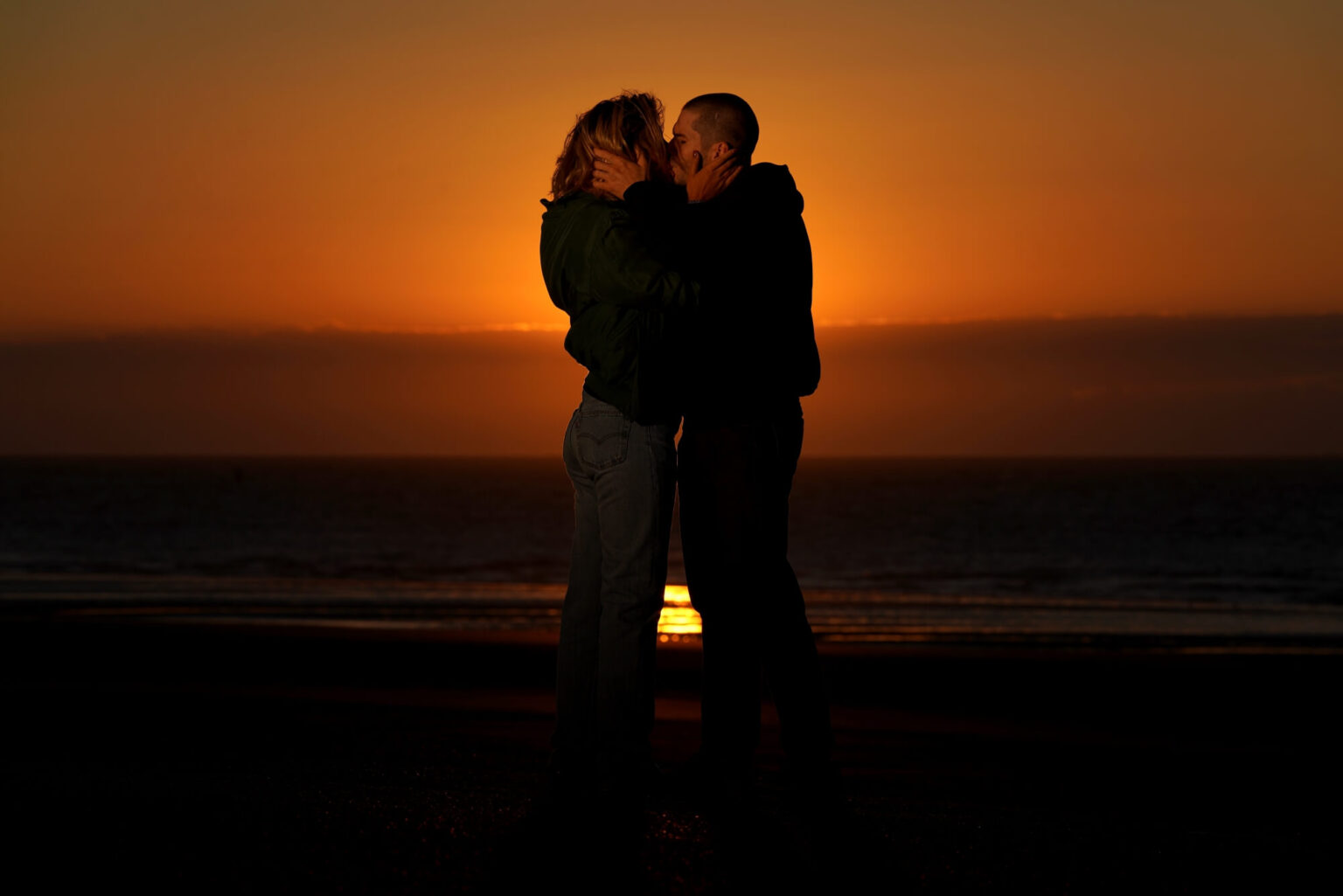 Para stoi blisko siebie na plaży o zachodzie słońca, ich twarze znajdują się blisko siebie, słońce jest nisko nad horyzontem, a w tle widać ocean.