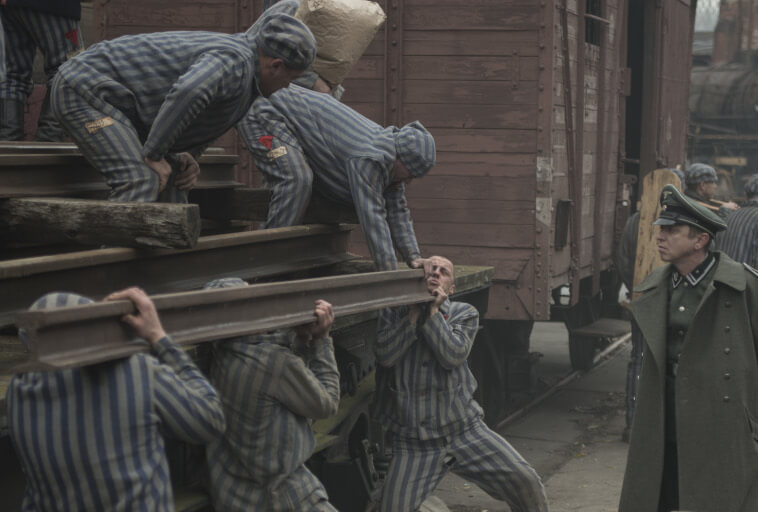 Kilku więźniów w pasiakach walczy o podniesienie ciężkich metalowych belek w pobliżu pociągu, podczas gdy obserwuje ich umundurowany strażnik w zielonym płaszczu i czapce. Scena wydaje się ponura i napięta.
