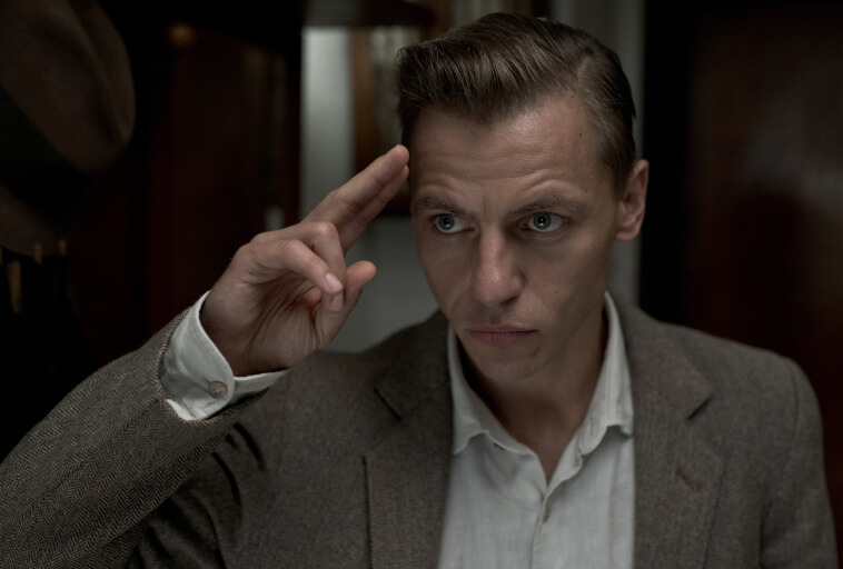 Mężczyzna w jasnej koszuli i brązowej marynarce podnosi rękę do czoła w geście pozdrowienia, wyglądając na poważnego i skupionego. Tło jest słabo oświetlone z widocznymi drewnianymi meblami.