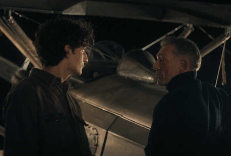 Dwaj mężczyźni stoją nocą obok staromodnego dwupłatowca i rozmawiają ze sobą. Przyćmione oświetlenie tworzy dramatyczną atmosferę, podkreślając zabytkowy samolot za nimi.