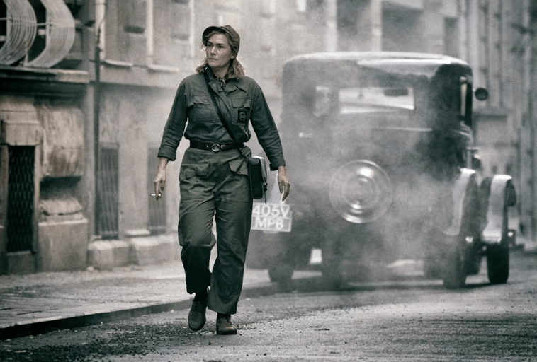 Kobieta w wojskowym stroju idzie opustoszałą, zadymioną ulicą obok staromodnego samochodu, ze zniszczonymi budynkami w tle, przywołując wojenną scenerię.