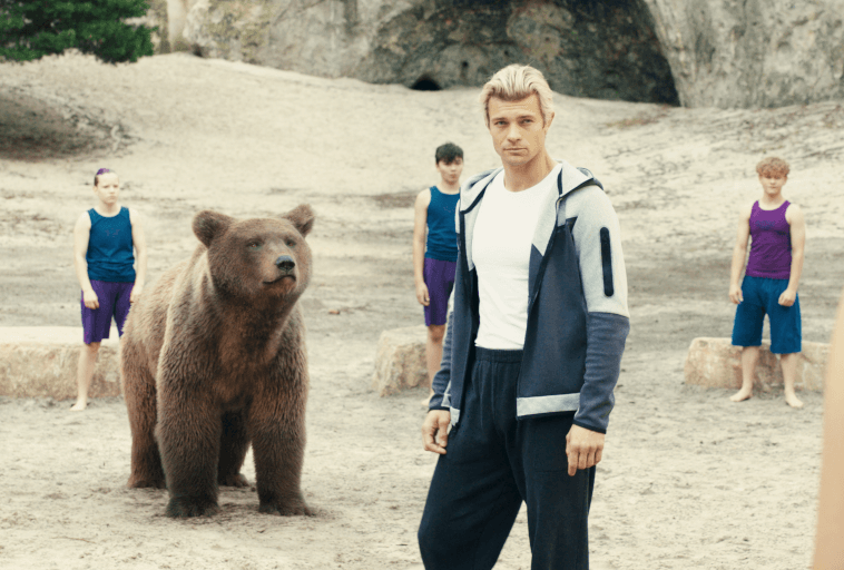 Mężczyzna w sportowym stroju stoi na piaszczystym terenie obok niedźwiedzia, a w tle stoją cztery osoby w pasujących fioletowych i niebieskich strojach sportowych, wszyscy na zewnątrz z formacjami skalnymi za nimi.