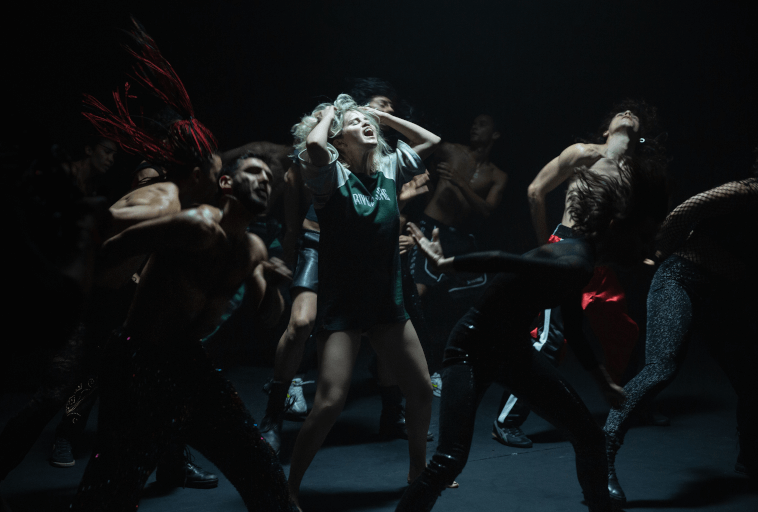 Grupa tancerzy w dramatycznych pozach występuje energicznie na ciemnej scenie, z blondynką w centrum chwytającą się za głowę, a inni pochylają się i poruszają ekspresyjnie wokół niej.