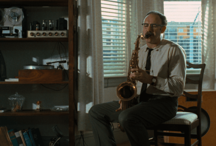 Mężczyzna w okularach i z wąsami siedzi na krześle w oświetlonym słońcem pokoju i gra na saksofonie. Na półkach obok leżą książki, sprzęt audio i gramofon. Światło wpada przez żaluzje okienne za nim.
