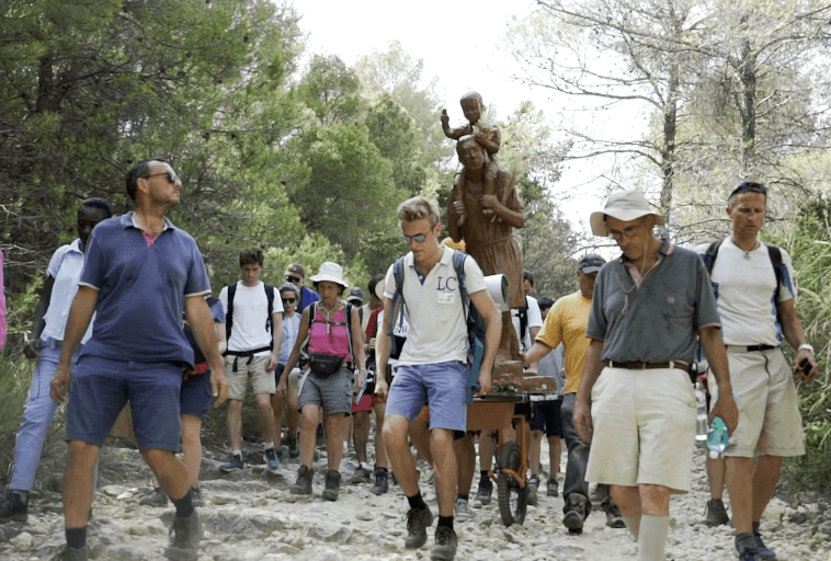 Grupa wędrowców idzie skalistym leśnym szlakiem, otaczając i niosąc posąg postaci w szacie z dzieckiem. Grupa jest ubrana w swobodną odzież outdoorową i otoczona drzewami.