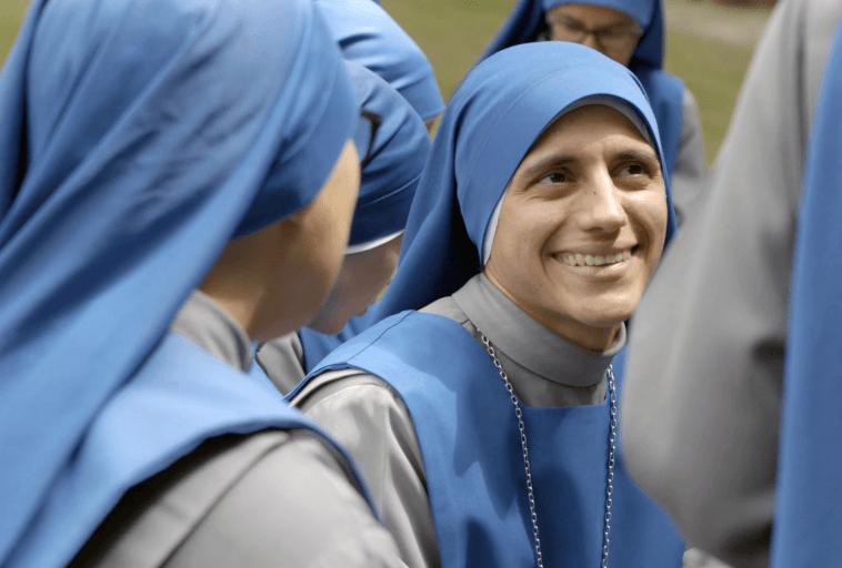 Uśmiechnięta zakonnica w niebiesko-szarym habicie siedzi wśród innych zakonnic ubranych podobnie, prowadząc rozmowę na świeżym powietrzu.