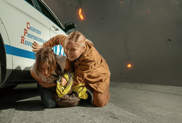 Dwie kobiety przykucają obok radiowozu, aby się osłonić, podczas gdy w tle dochodzi do eksplozji, a w powietrzu widoczne są szczątki i płomienie. Jedna z kobiet osłania drugą.