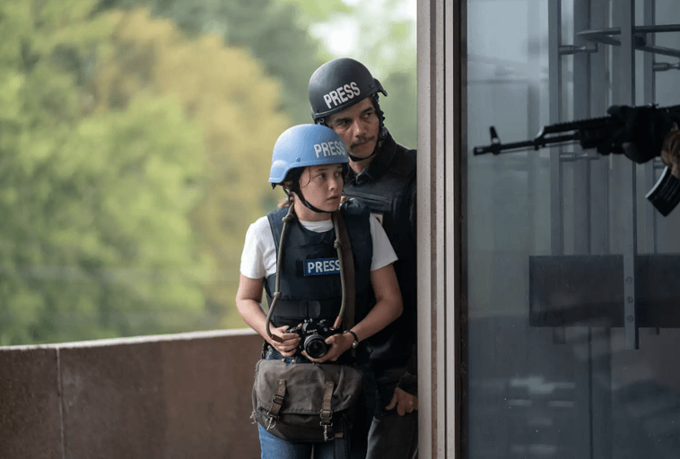 Dwóch dziennikarzy w kaskach PRESS i kamizelkach kuloodpornych ostrożnie stoi za ścianą. Jeden trzyma kamerę, drugi wygląda na czujnego. Z wnętrza budynku widać pistolet uzbrojonej osoby skierowany w ich stronę.