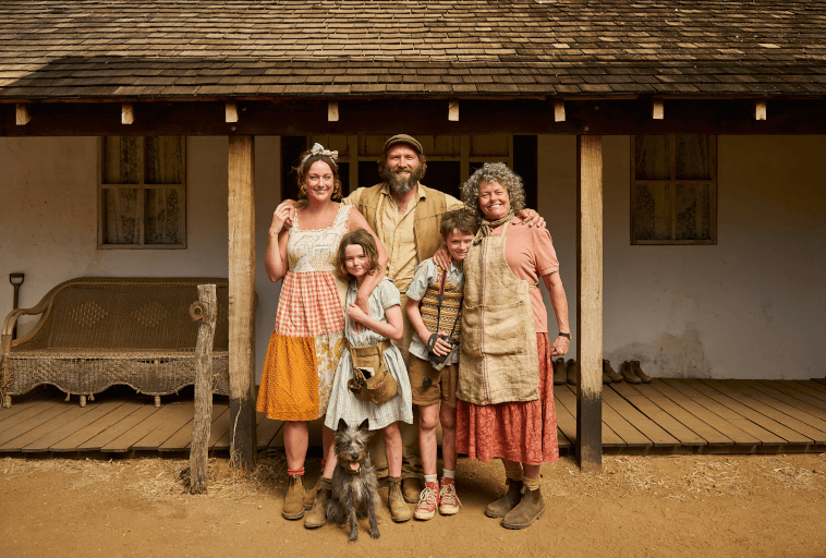 Uśmiechnięta pięcioosobowa rodzina stoi na ganku przed rustykalnym domem, ubrana w wiejskie ubrania w stylu vintage. Dwoje dorosłych, dwoje dzieci, starsza kobieta i mały pies pozują razem.