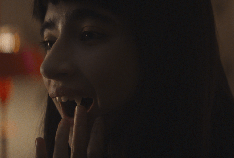 Osoba z długimi ciemnymi włosami trzyma palce przy ustach, pokazując sztuczne wampirze kły, patrząc w bok w miękkim, słabym oświetleniu.