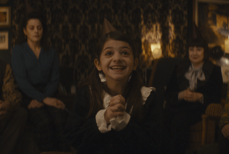 Młoda dziewczyna z warkoczami i imprezowym kapeluszem uśmiecha się podekscytowana, ściskając dłonie, siedząc w ciepło oświetlonym pokoju z dorosłymi w tle, prawdopodobnie podczas uroczystości.