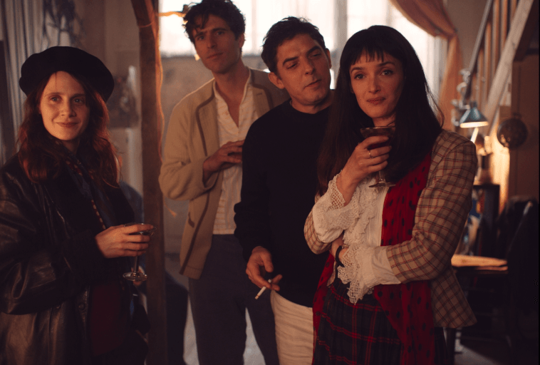 Cztery osoby stoją w pomieszczeniu na spotkaniu towarzyskim, trzymając drinki. Są ubrani w stylowe ubrania inspirowane stylem vintage i wydają się cieszyć swoim towarzystwem. Ciepłe oświetlenie tworzy przytulną atmosferę.