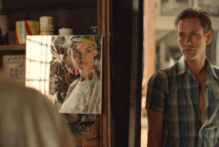 Mężczyzna w kraciastej koszuli stoi w pomieszczeniu obok lustra z popękaną powierzchnią, która odbija twarz kobiety noszącej kolorową chustę. W tle widać półki z różnymi przedmiotami.