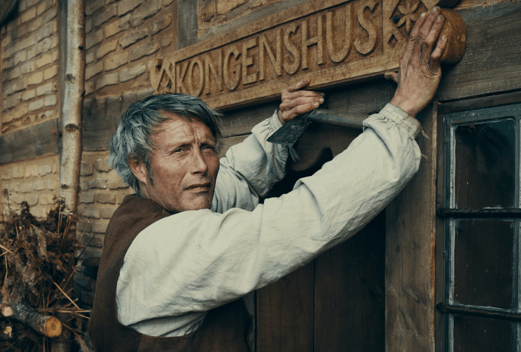 Mężczyzna z siwiejącymi włosami i w staromodnym ubraniu instaluje drewnianą tabliczkę z napisem KONGENSHUS nad rustykalnymi drzwiami ceglanego budynku, trzymając w jednej ręce narzędzie.