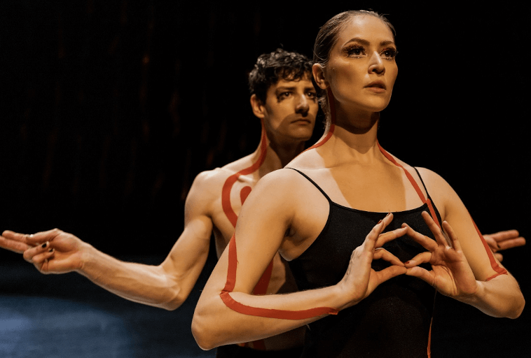 Dwoje tancerzy stoi w dramatycznej pozie na scenie. Kobieta z przodu ma na sobie czarny strój i wykonuje gest rękami, podczas gdy stojący za nią mężczyzna bez koszulki wyciąga ramiona, oboje z czerwonymi liniami namalowanymi na ich ciałach.