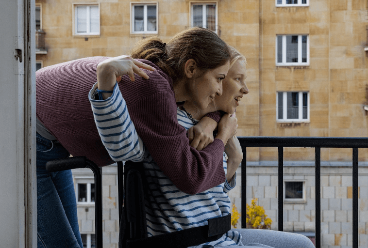 Dwie młode kobiety, jedna siedząca na wózku inwalidzkim, a druga stojąca za nią, uśmiechają się i obejmują, patrząc razem na balkon. W tle widoczny jest budynek mieszkalny.