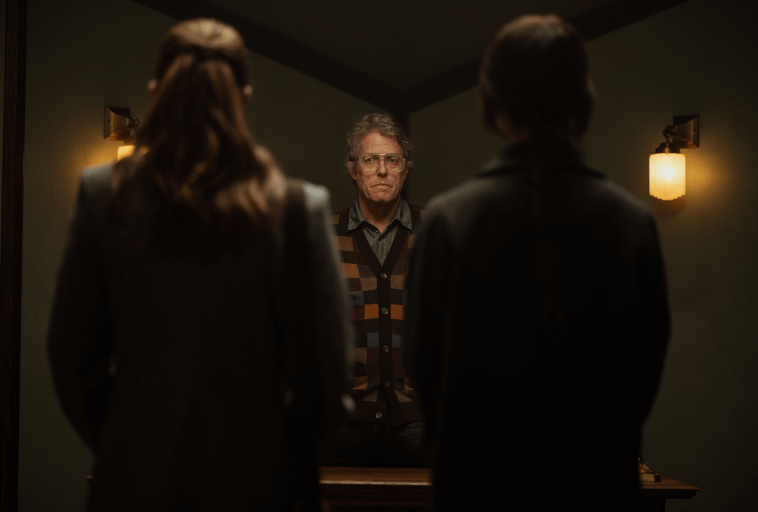 Mężczyzna w okularach i wzorzystym swetrze stoi naprzeciwko dwóch osób z długimi włosami, widzianych od tyłu, w słabo oświetlonym pokoju z zielonymi ścianami i dwoma kinkietami.