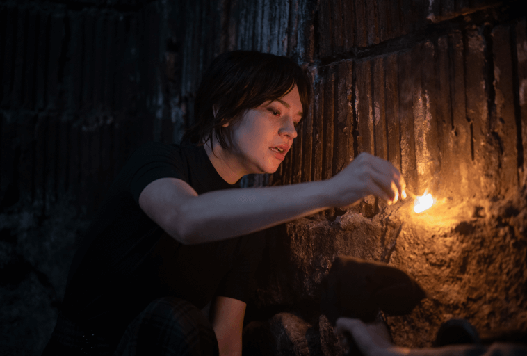 Osoba o krótkich ciemnych włosach zapala zapałkę na szorstkiej, ciemnej kamiennej ścianie, oświetlając swoją twarz w słabym, zacienionym otoczeniu.