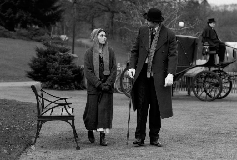Kobieta w staromodnym ubraniu idzie obok mężczyzny w płaszczu, kapeluszu i rękawiczkach trzymającego laskę. Znajdują się w parku w pobliżu ławki, z powozem konnym i woźnicą w tle. Obraz jest czarno-biały.