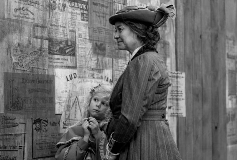 Czarno-białe zdjęcie kobiety w historycznym stroju i kapeluszu stojącej obok młodej dziewczyny o jasnych włosach, która patrzy na nią. Oboje stoją przed drewnianą ścianą pokrytą starymi plakatami i ogłoszeniami.