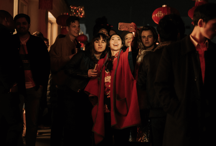 Grupa ludzi stoi blisko siebie w nocy, niektórzy się uśmiechają. Dwie kobiety w czerwonych odświętnych ubraniach robią sobie selfie, podczas gdy inni obserwują lub rozmawiają. Czerwone latarnie wiszą nad nimi, tworząc ciepłą, świąteczną atmosferę.