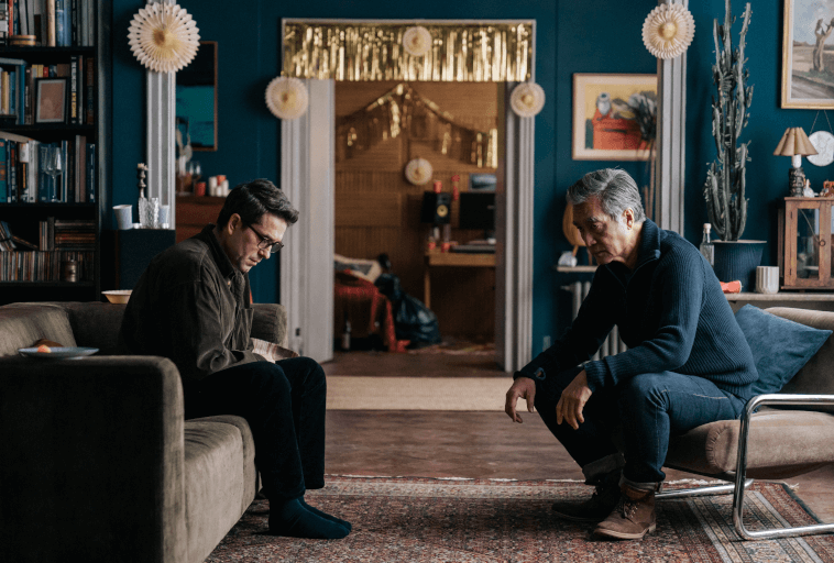 Dwóch mężczyzn siedzi naprzeciwko siebie na oddzielnych kanapach w przytulnym, ciepło oświetlonym salonie, obaj wyglądają na poważnych i głęboko zamyślonych. Półki z książkami, dzieła sztuki i dekoracje wypełniają stylową przestrzeń wokół nich.