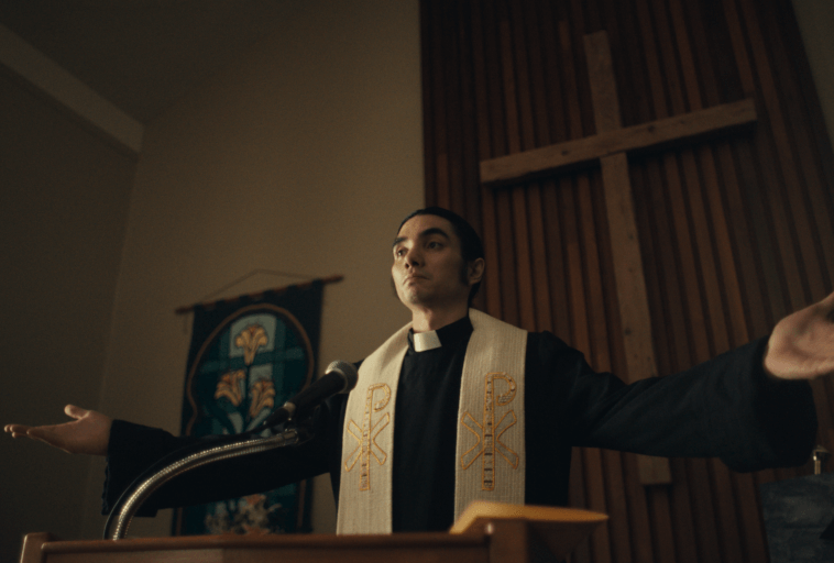 Ksiądz w czarnych szatach i białej stule stoi na kościelnej ambonie z wyciągniętymi rękami, za mikrofonem, przed dużym drewnianym krzyżem i ozdobnym gobelinem.