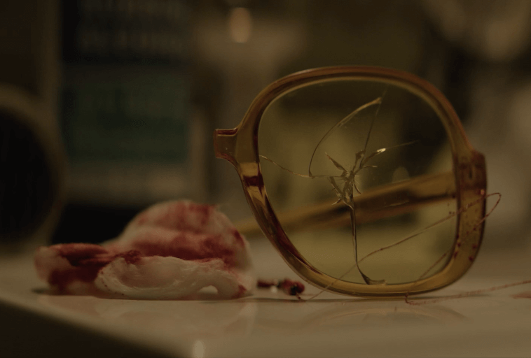 Para żółtych okularów z pękniętą soczewką leży na powierzchni obok zakrwawionego wacika, co sugeruje uraz lub gwałtowny incydent. Tło jest rozmyte.