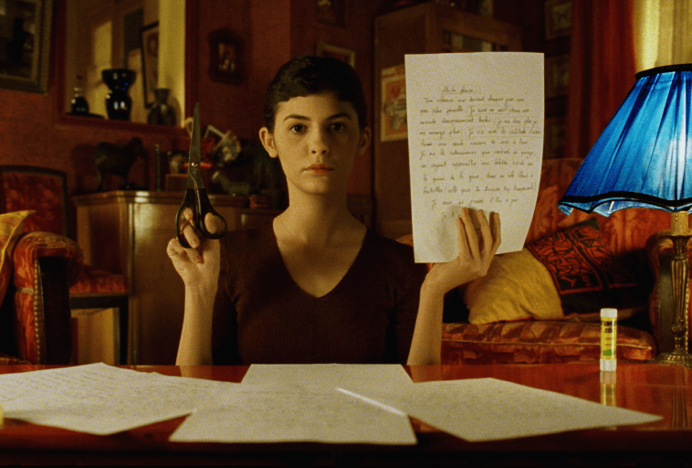 Kobieta o krótkich ciemnych włosach siedzi przy stole, trzymając w jednej ręce nożyczki, a w drugiej odręcznie napisany list. Patrzy bezpośrednio w kamerę. Pokój jest ciepło oświetlony w odcieniach czerwieni i pomarańczy.