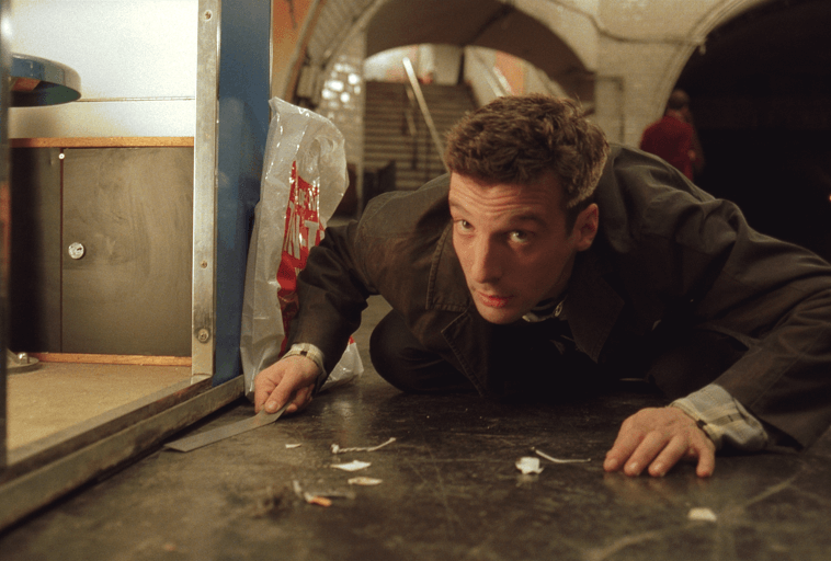 Mężczyzna w ciemnym płaszczu przykuca nisko na podłodze stacji metra, patrząc uważnie w górę, ze śmieciami rozrzuconymi wokół i schodami widocznymi w tle.