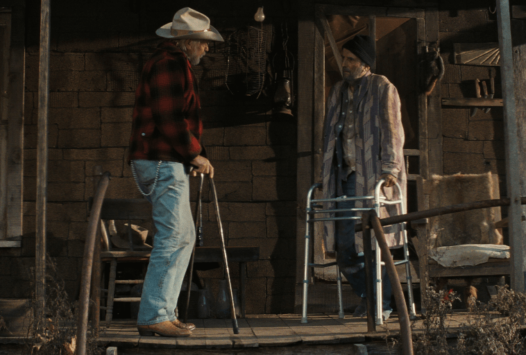 Dwóch starszych mężczyzn stoi na rustykalnej drewnianej werandzie. Jeden nosi kowbojski kapelusz i używa laski, podczas gdy drugi, w czapce i pasiastym szlafroku, korzysta z chodzika. Ganek wygląda na zużyty, a na ścianie wiszą skóry zwierząt.