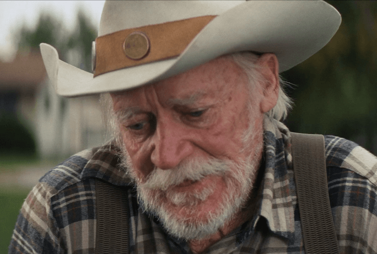 Starszy mężczyzna z białą brodą i wąsami w kowbojskim kapeluszu i koszuli w kratę spogląda w dół z zamyślonym wyrazem twarzy. Tło jest rozmyte, co sugeruje ustawienie na zewnątrz.