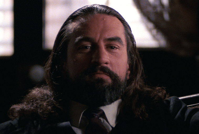 Mężczyzna z długimi ciemnymi włosami i pełną brodą, ubrany w garnitur i krawat, siedzi w słabo oświetlonym pokoju, patrząc prosto w kamerę z poważnym wyrazem twarzy.